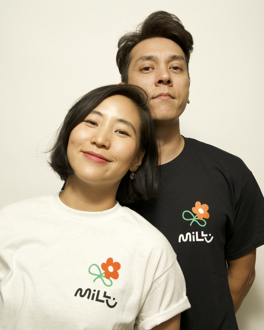 Two people wearing white and black MILLU Bloom T-shirts, showing the orange flower and MILLU logo on the chest.