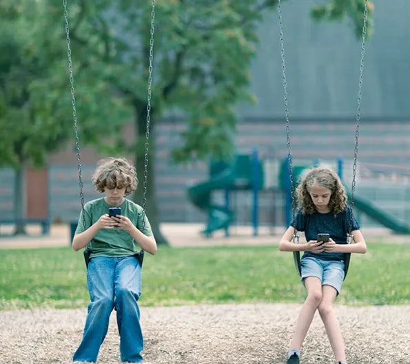 two kids on swings looking on their phones