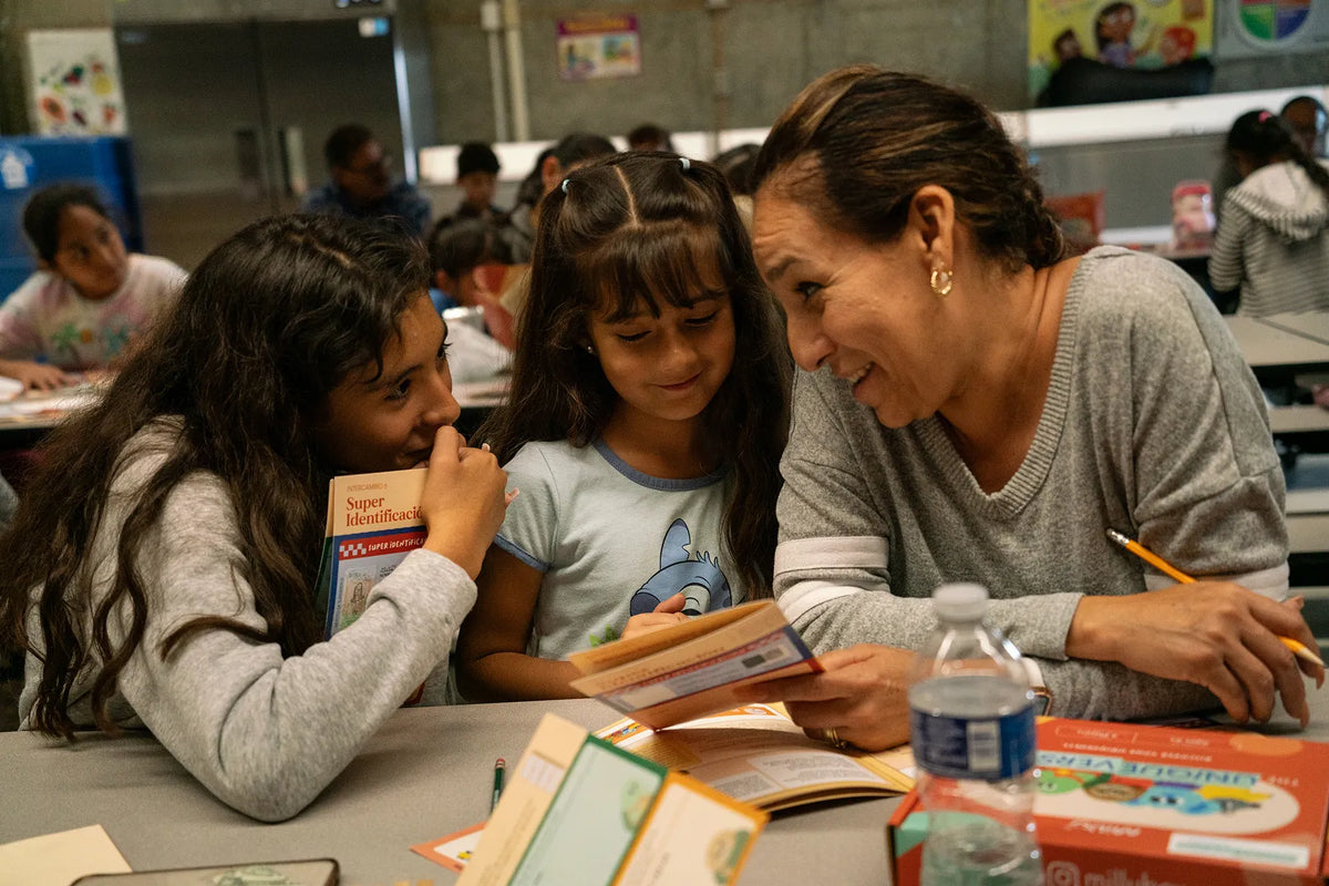 Woman and two young girls sitting at a table reading books together in a classroom setting.