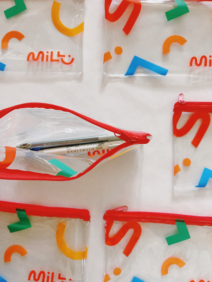 Open clear MILLU pencil case showing pens inside, with a red zipper and colorful geometric print.